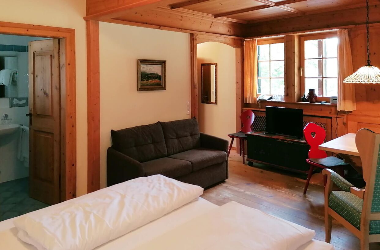 Berghotel Sudelfeld Bayrischzell Ferienwohnung in einem Nebengebäude mit Laminatboden; im Vordergrund ein Doppelbett, dahinter Schlafsofa, Holztisch, ein Hochstuhl, ein kleiner roten Sessel und Deckenlampe