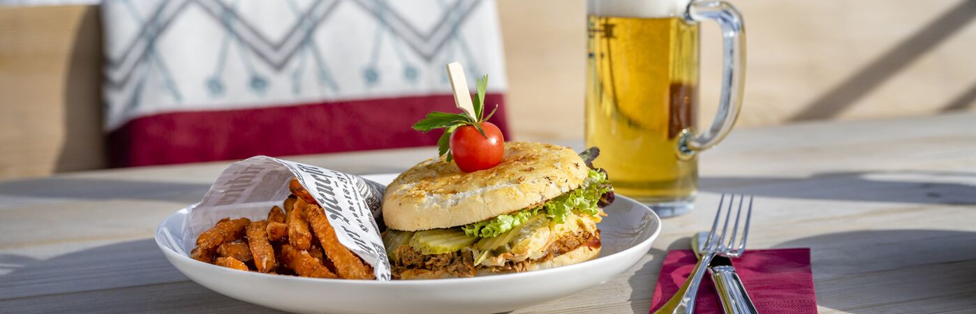 Restaurant Broesel Alm auf einem Holztisch auf der Sonnenterrasse steht ein Teller mit Broesel Alm Burger Suesskartoffel-Pommes Besteck mit Serviette und ein Bier im Glaskrug