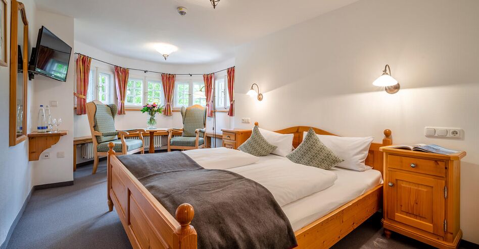 Berghotel Sudelfeld Bayrischzell Doppelzimmer mit gemuetlichem Erker Zimmer mit Doppelbett rustikale Einrichtung im Erker runder Tisch Blumenstrauss zwei Ohrensessel TV an der Wand 