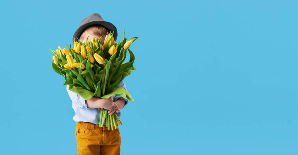 Ein kleiner Bub mit Trachtenhut und einem großen Strauß gelber Tulpen vor einem strahlend blauen Himmel